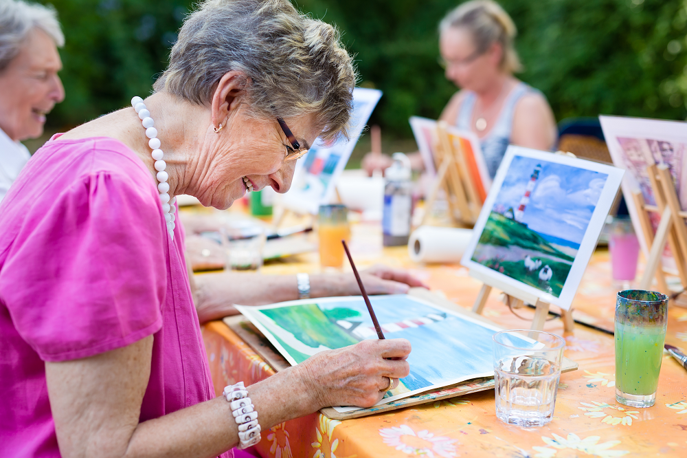 An older woman sits at a table and paints.
